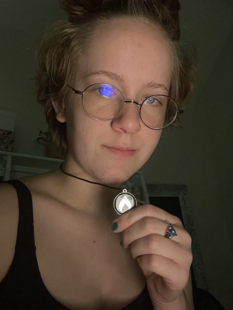 A closeup photo of me, a white teenager with auburn hair in a messy topknot. I am wearing wire-frame glasses and there is a blue glare on the left lense. I am wearing a black tank top and holding a necklace with a black pendant with a silver chalice on it.